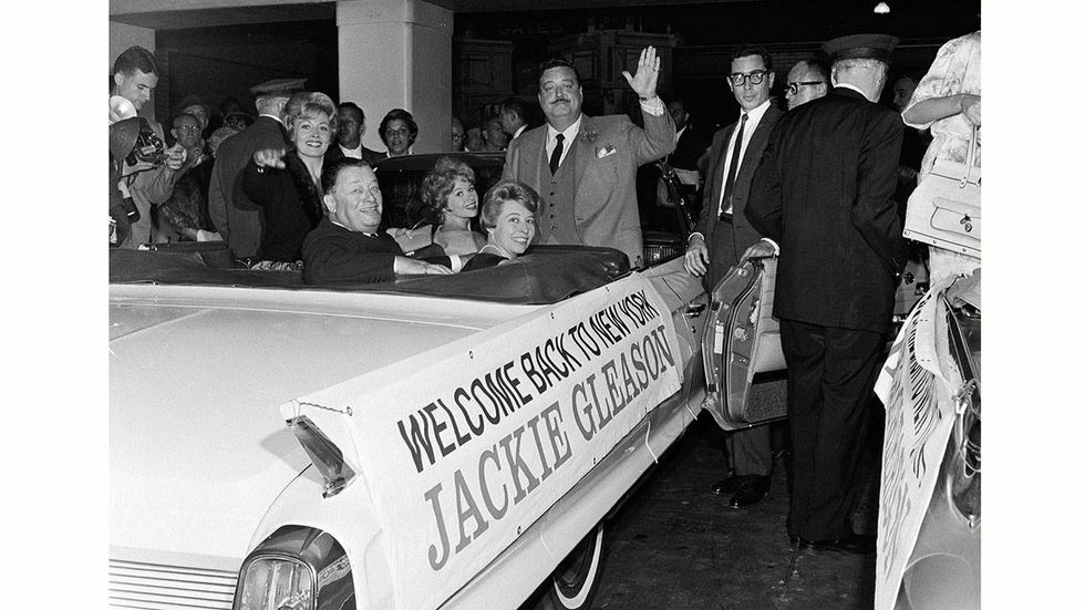 American actor and entertainer Jackie Gleason (1916-1987) stands in a Cadillac and waves upon his return to his hometown, outside Penn Station, New York, New York, August 18, 1962.  Smiling from car's backseat is American restauranteur Bernard Toots Shor (1904-1977).
